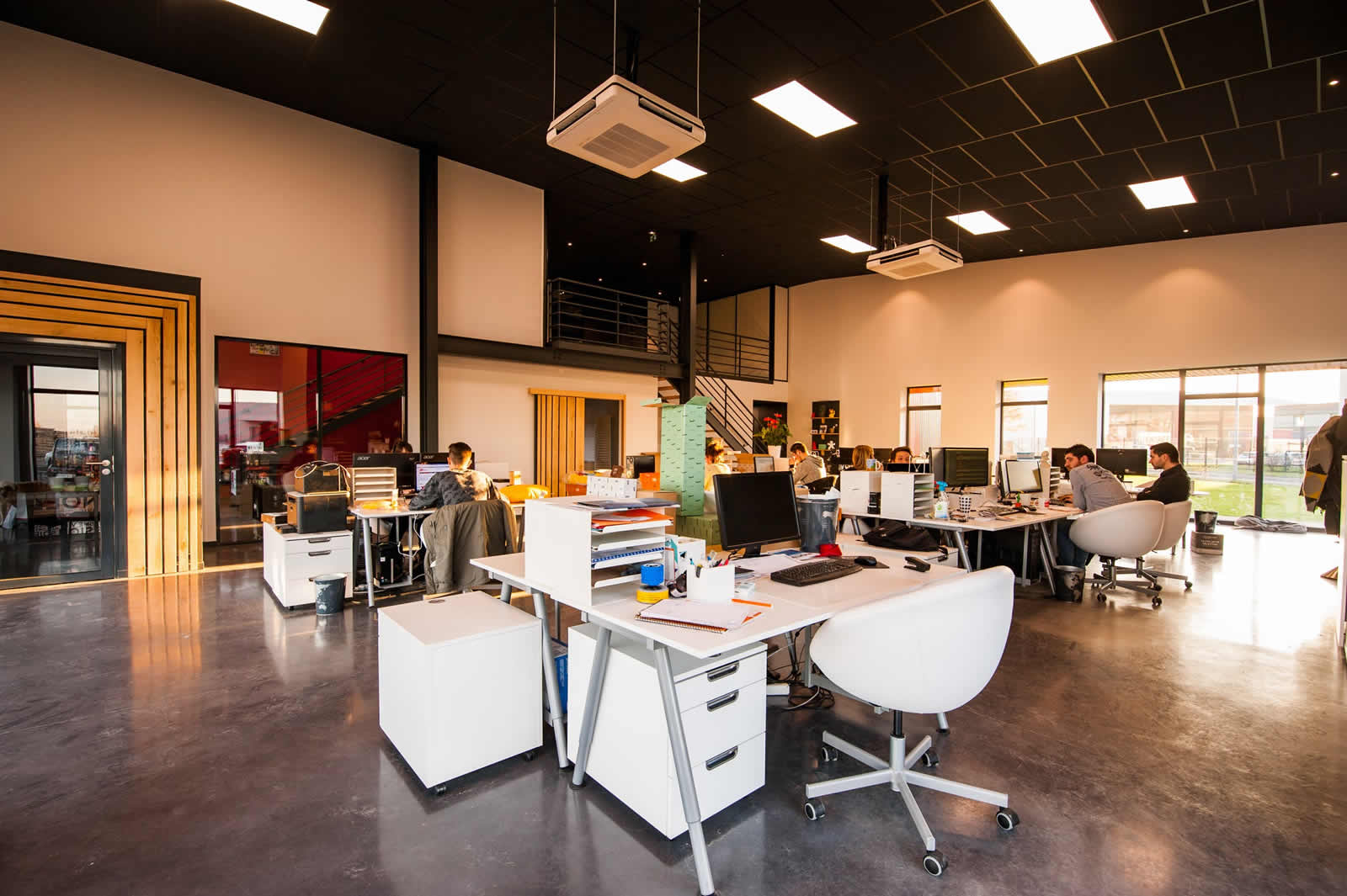 Modern open-plan office with employees working at shared desks, large windows, high ceilings, and contemporary lighting.