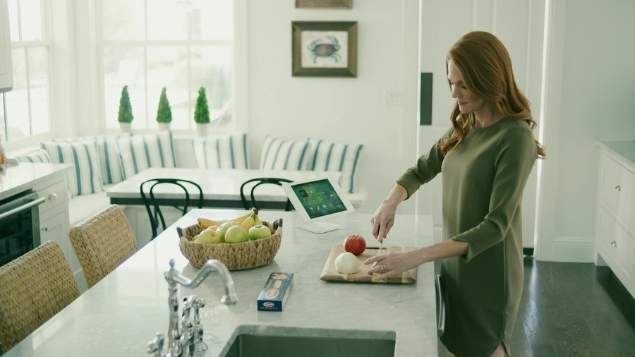 woman chopping vegetables at a kitchen island. A Control4 touchscreen is on the counter beside her.