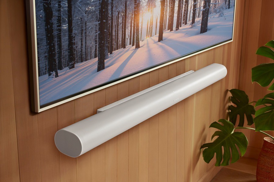 Sonos’ Arc Ultra soundbar in white, mounted beneath a wall-mounted TV displaying a snowy forest scene, set against a wooden paneled wall with a lush green plant nearby.