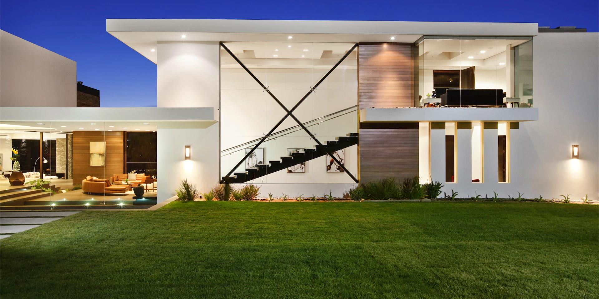 A modern two-story house with a large glass facade, sleek exterior design, and a well-manicured lawn, illuminated beautifully in the evening.
