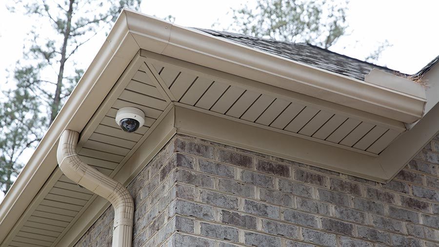 An outdoor security camera mounted under the soffit of a house, with a brick exterior and a view of nearby trees.