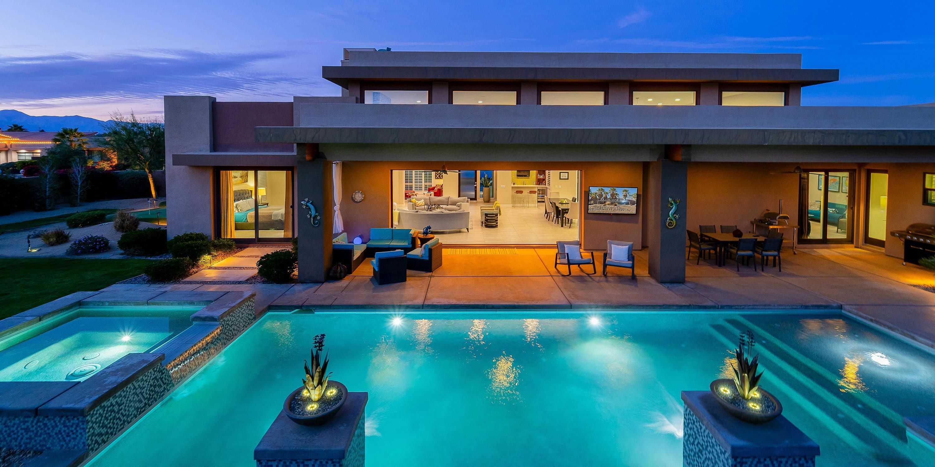 An illuminated outdoor pool area with a connected spa, surrounded by modern patio seating and an open indoor living space at dusk.