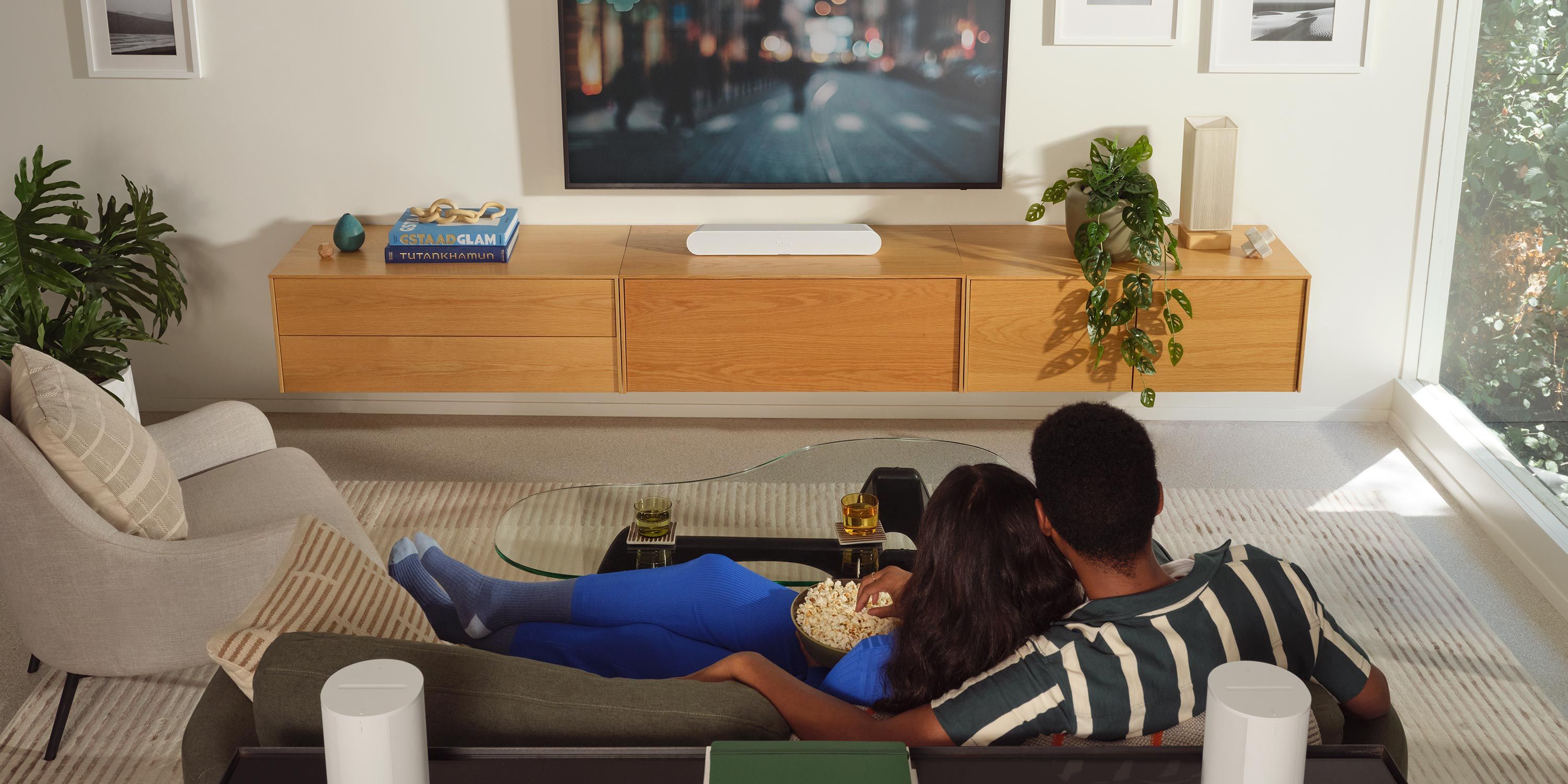 A cozy media room with a couple sitting on a sofa, watching a large wall-mounted TV, with a modern console and decorative plants.